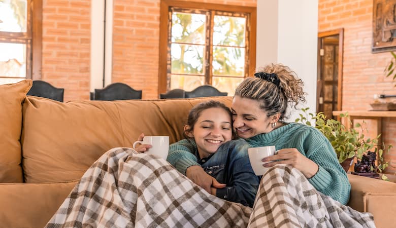 Mãe e filha embaixo de cobertas, sentadas no sofá e tomando um chá, recuperando de problemas respiratórios no inverno.