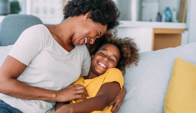Mãe e filha abraçadas e sorrindo no sofá, em referência a respirar bem. 