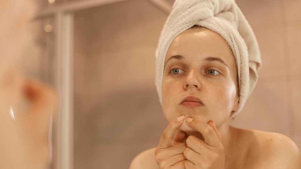 Jovem com uma toalha enrolada na cabeça olha-se no espelho, usando os dedos para pressionar uma espinha ou acne na região do queixo
