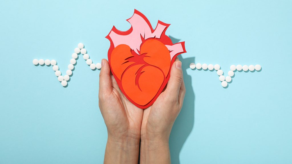 Duas mãos seguram um recorte de papel em formato de coração humano contra um fundo azul-claro, com linha pontilhada branca formando linha de ritmo cardíaco de eletrocardiograma (ECG).