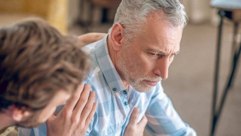 Homem mais novo coloca a mão no ombro de um homem mais velho de cabelos grisalhos com a mão no peito, na altura do coração, com expressão preocupada.