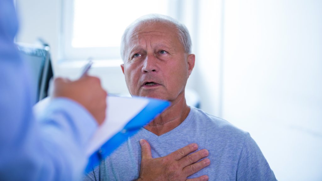 Homem mais velho com expressão preocupada, sentado em um ambiente médico, segura a mão contra o peito enquanto olha para um profissional de saúde que faz anotações.