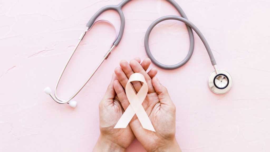 Mãos segurando um laço e um estetoscópio sobre um fundo rosa, simbolizando a conscientização sobre o câncer e a importância do check-up de prevenção