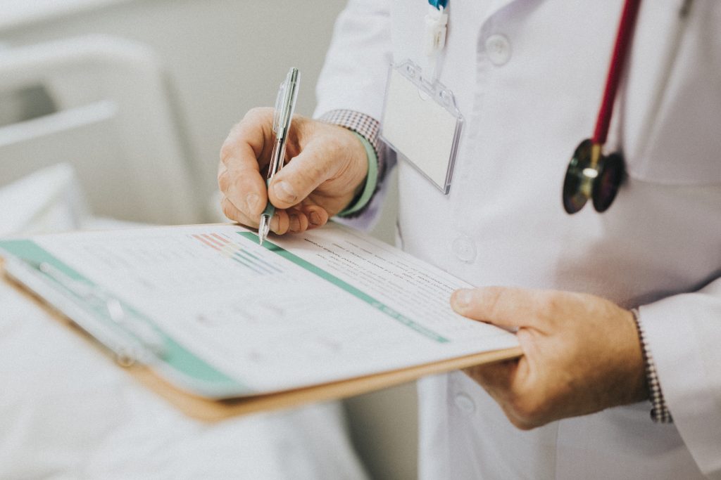Médico de jaleco branco e estetoscópio no pescoço segura uma prancheta e usa caneta para anotar dados em uma ficha médica em um hospital