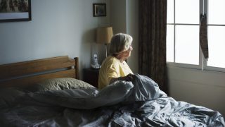mulher idosa sentada em uma cama e olhando para a janela do quarto com atitude que retrata um quadro de depressão em pessoas da terceira idade