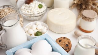 Diversos laticínios, ovos, um biscoito com gotas de chocolate, aveia e talos de trigo estão dispostos sobre uma superfície branca, exemplificando alimentos a serem evitados por quem sofre de intolerância à lactose ou de alergia ao leite