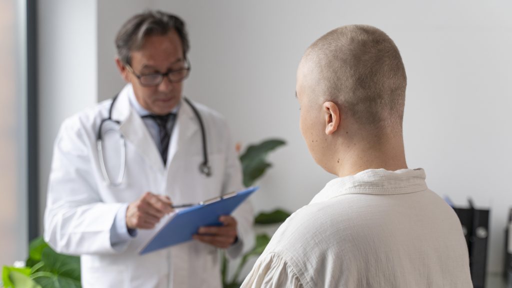 Um médico de jaleco branco e estetoscópio no pescoço segura uma prancheta azul enquanto está diante de um paciente de cabeça raspada em uma clínica