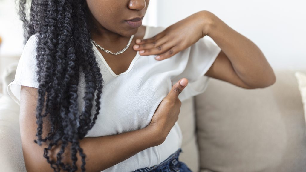 Mulher de cabelos escuros, longos e trançados, vestindo blusa branca com decote em V, realiza um autoexame nas mamas posicionando as mãos sobre o peito enquanto está sentada.