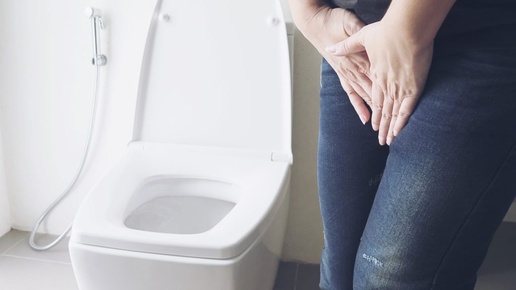 mulher com as mãos na virilha ao lado de um vaso sanitário, fazendo referência à incontinência urinária durante a menopausa