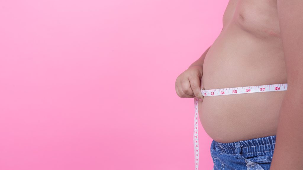 Homem com sobrepeso está e pé em frente a um fundo rosa e mede sua barriga usando uma fita métrica