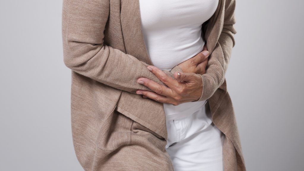 Mulher vestindo um cardigã bege e uma blusa branca segura a barriga, indicando dor e desconforto provocados pelas cólicas menstruais