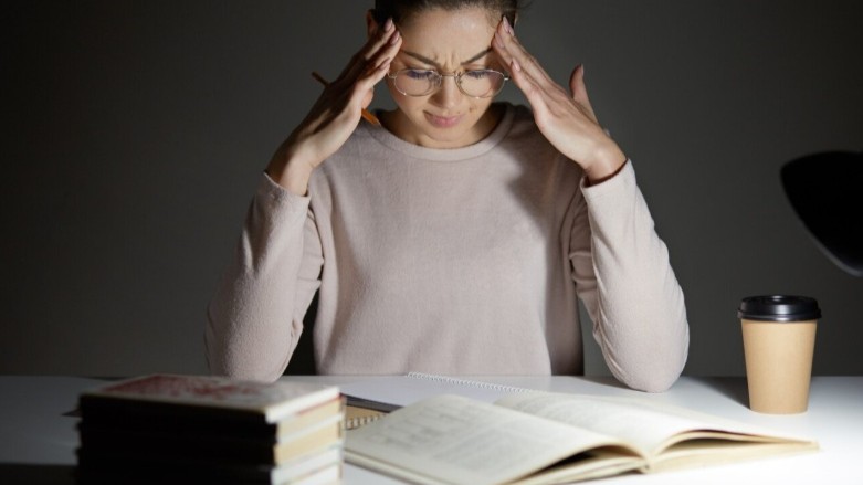 mulher jovem sentada diante de uma mesa com vários livros e um copo de café, com expressão angustiada e as mãos apoiadas na cabeça, representando a dificuldade de concentração de uma pessoa com TDAH