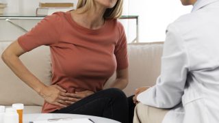 mulher sentada em um consultório médico, com as mãos no abdômen sinalizando dor provocada por infecção pela bactéria H. pylori; à sua frente está uma médica vestindo jaleco branco