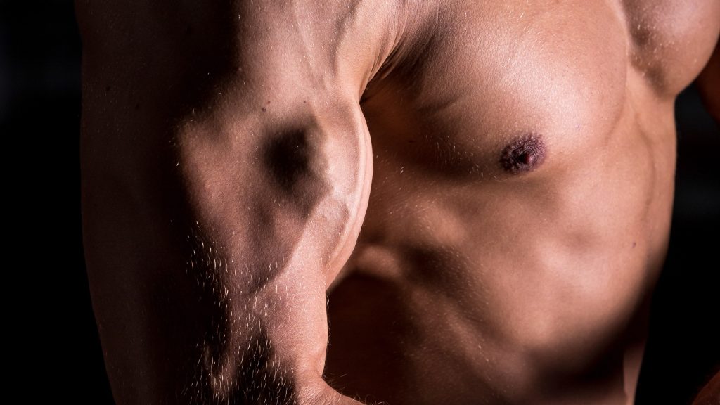 Tronco e braço de um homem musculoso, destacando a definição dos músculos do bíceps, do peitoral e do abdômen contra um fundo escuro para ilustrar efeitos do uso de esteroides anabolizantes